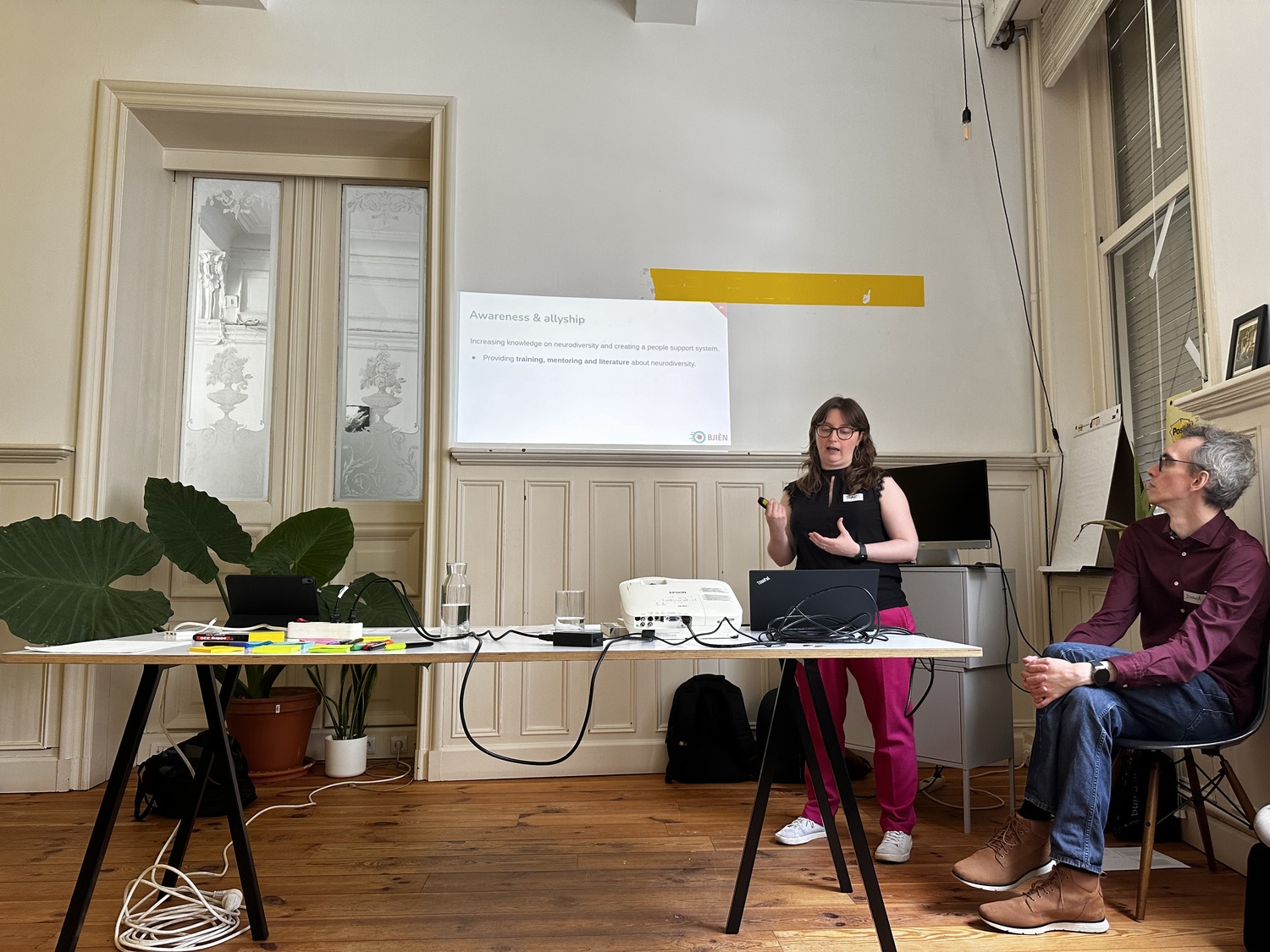 Daphné from Bjièn standing behind a table and presenting. Behind her, a slide with the title 'Awareness & allyship' is visible. Her colleague Dietrich sits next to her and looks at the slide.