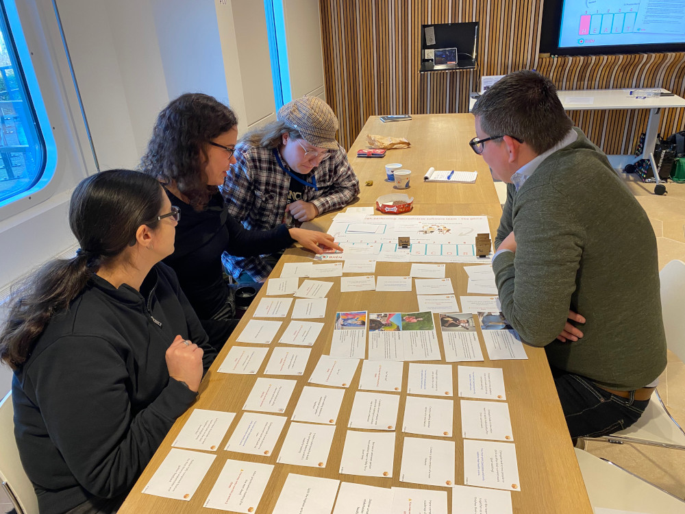 A group of four participants playing the board game while seated at a table. On the table the playing board is visible, along with a bunch of practices cards. Participants are looking closely at the cards.