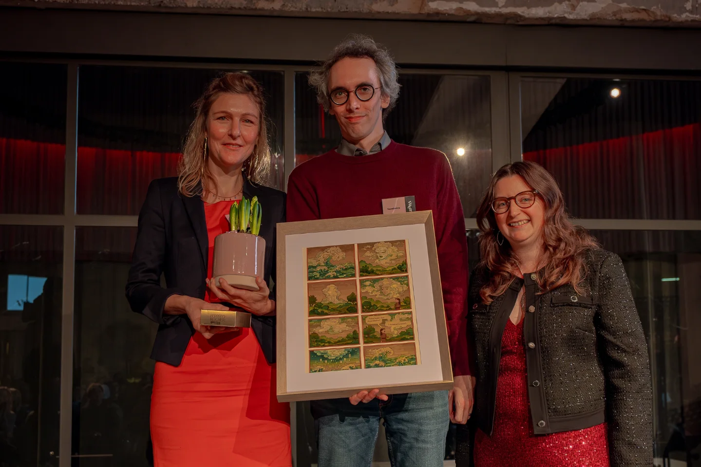 Marjan, Dietrich et Daphné sourient ensemble sur la photo. Dietrich tient le prix, une illustration encadrée. Marjan tient un pot de fleurs.