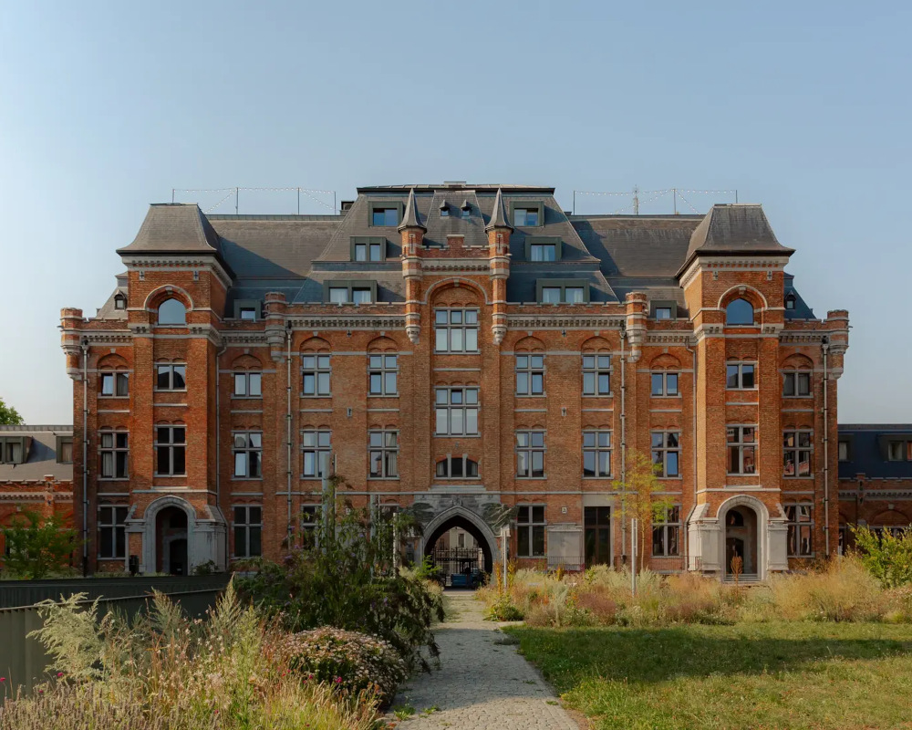 Groot historisch bakstenen gebouw met rijk versierde gevel, hoge ramen, torentjes en een leistenen dak, gezien vanaf een groene tuin met een wandelpad naar de centrale ingang.